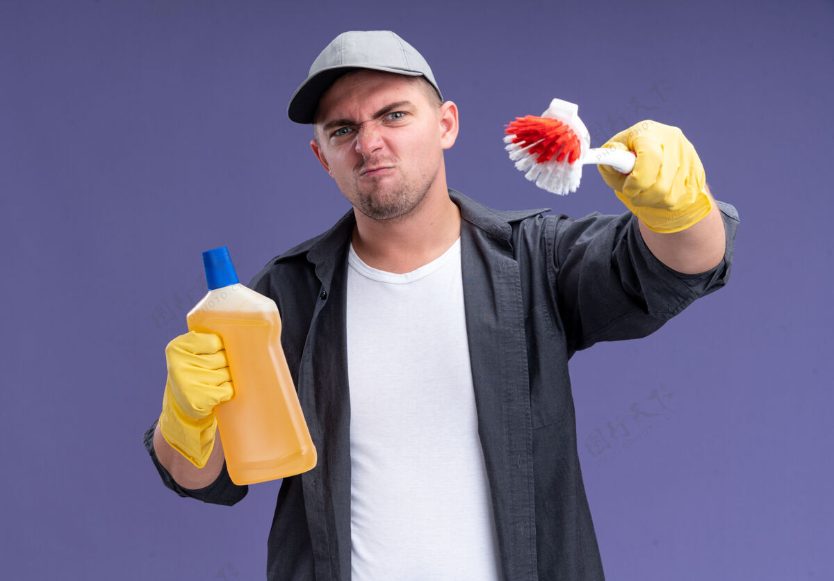 手套自信的年轻帅气的清洁工穿着t恤 戴着帽子和手套 手里拿着清洁剂 手里拿着刷子隔离在紫色的墙上帅气穿自信