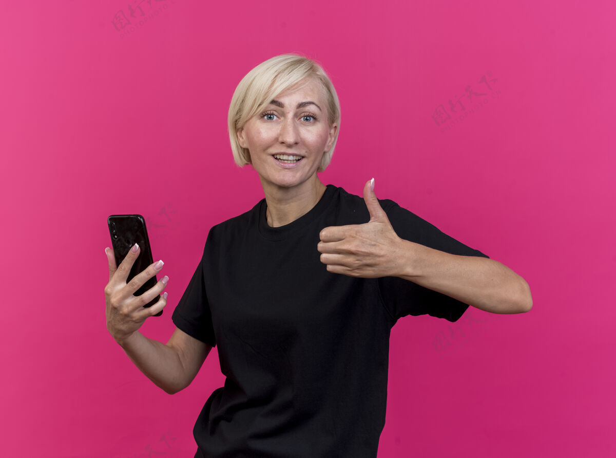 手机微笑着的中年金发斯拉夫女人拿着手机看着前面 在粉红色的墙上孤立地竖起大拇指姿势脸人