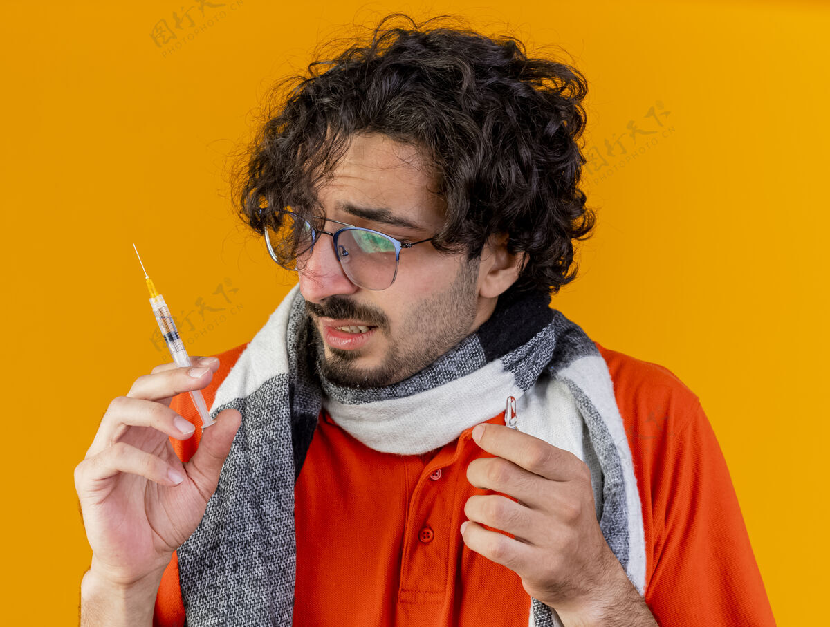 表情特写镜头：戴着眼镜 戴着围巾 拿着注射器和安瓿 看着隔离在橙色墙上的注射器的年轻病人衣服站着姿势