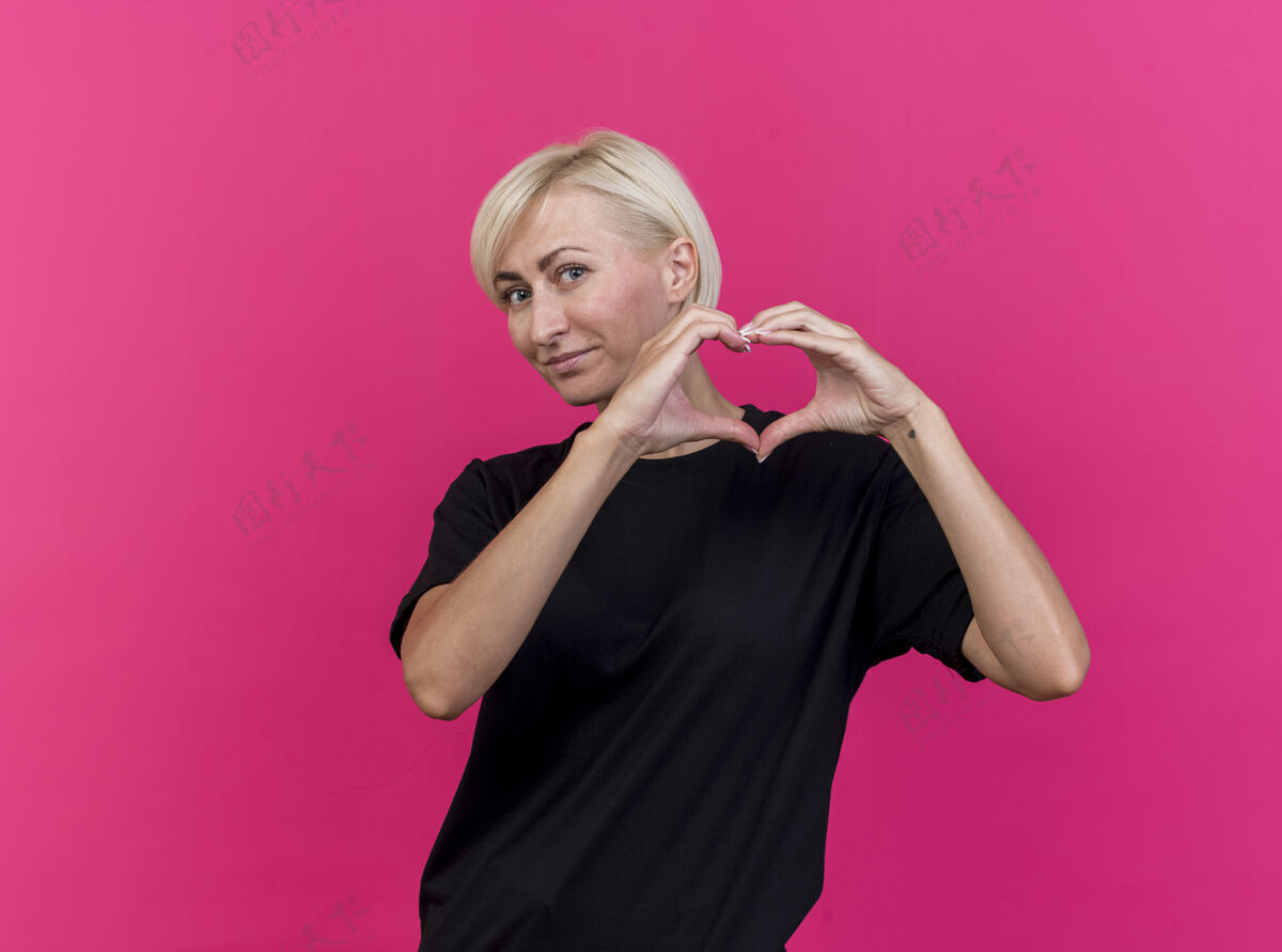 立场高兴的中年金发斯拉夫妇女看在前面做心脏标志孤立的粉红色墙上与复制空间衣服公民女人