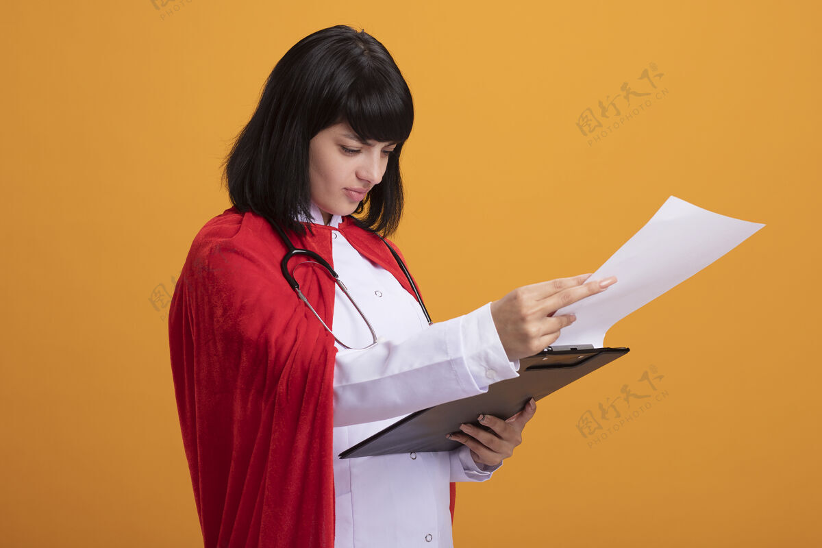 斗篷年轻的超级英雄女孩戴着听诊器 穿着医用长袍和斗篷 在橘色墙上的剪贴板上翻动医疗女孩剪贴板