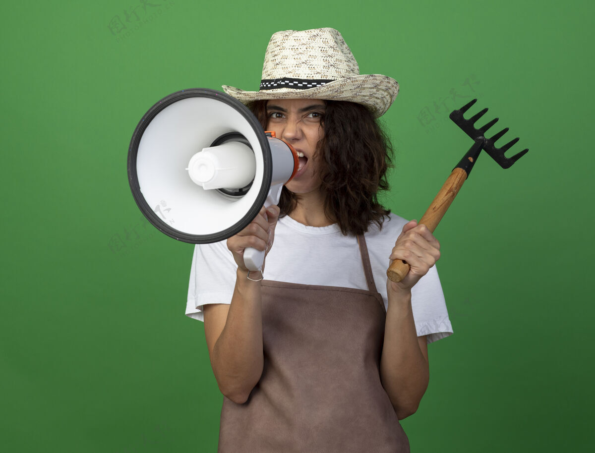 帽子愤怒的年轻女园丁穿着制服戴着园艺帽拿着耙子在扩音器上讲话花园扩音器女人