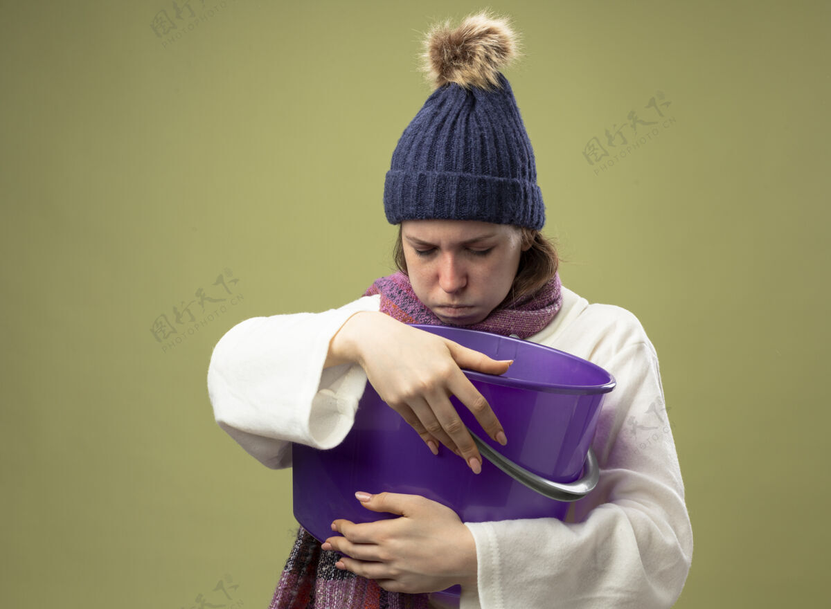 围巾虚弱的年轻生病的女孩穿着白色长袍 戴着冬天的帽子 戴着围巾 拿着塑料桶 恶心的感觉被隔离在橄榄绿上长袍虚弱桶
