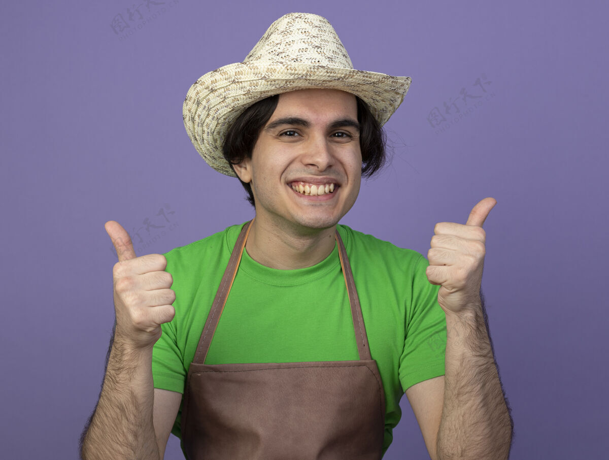 成功微笑着的年轻男园丁穿着制服 戴着园艺帽 竖起大拇指 孤立地站在紫色的地面上积极年轻