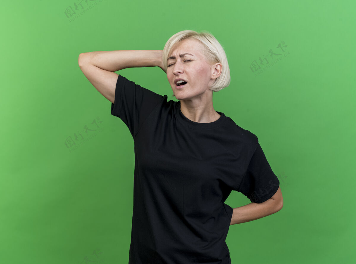 感觉疼痛的中年金发斯拉夫女人把双手放在头上和背上 闭着眼睛孤立在绿色的墙上衣服斯拉夫女人