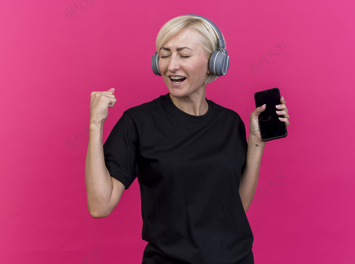 站着快乐的中年金发斯拉夫女人戴着耳机拿着手机做“是”的手势隔离在粉红色的墙上感觉女人金发