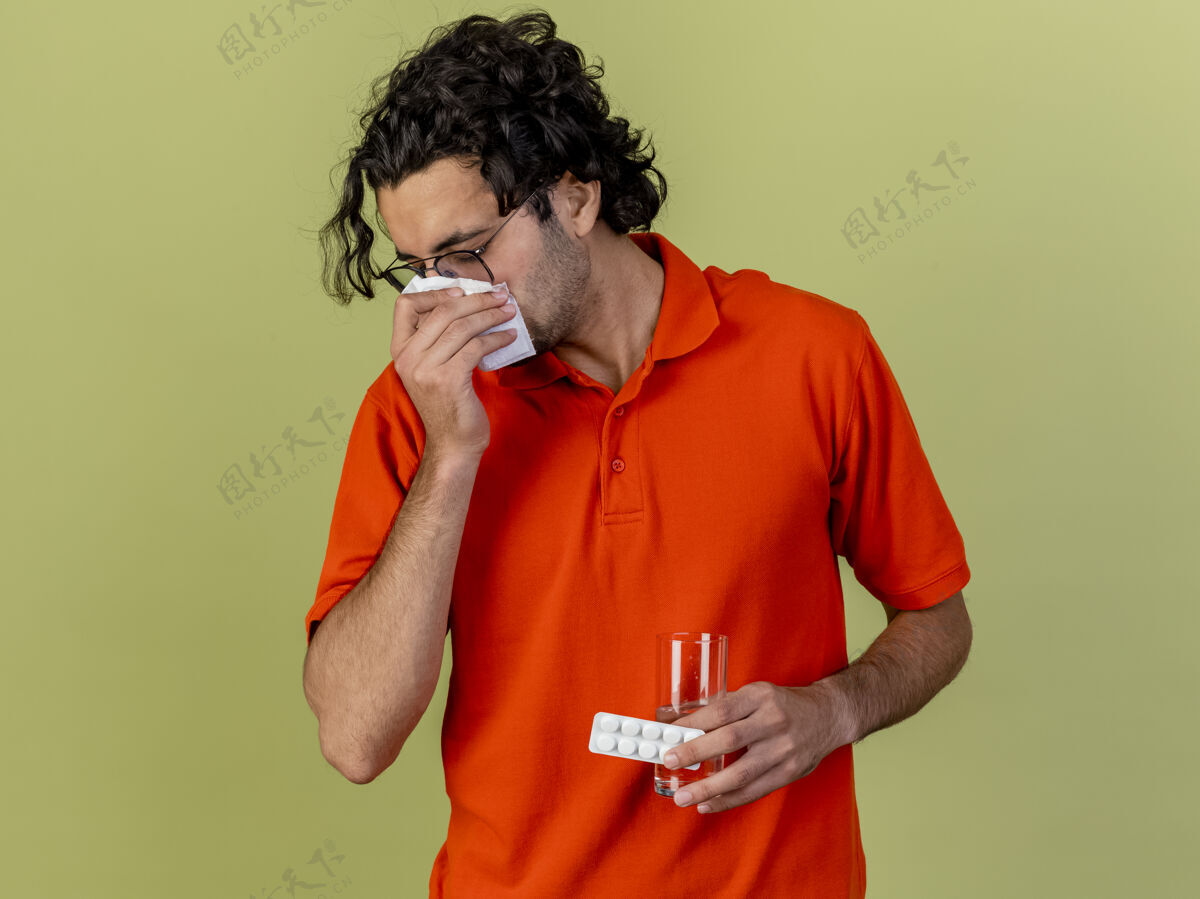 人年轻的病人戴着眼镜 拿着一杯水和一包药片 用餐巾擦鼻子 隔离在橄榄绿的墙上 留着复印空间衣服站着脸