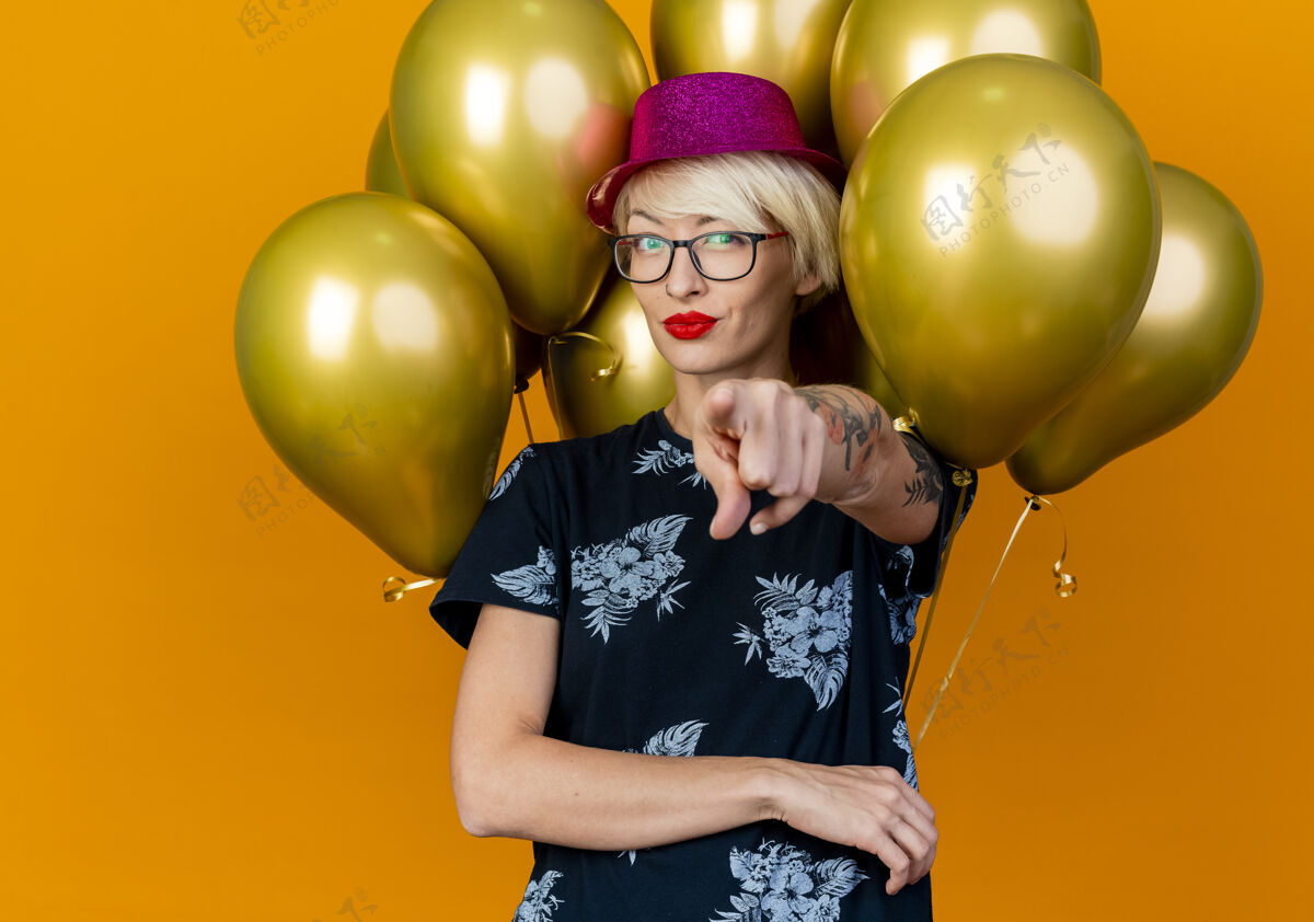 感觉自信的金发派对女郎戴着派对帽戴着眼镜站在气球前看着并指着橙色墙上孤立的前方衣服人前面