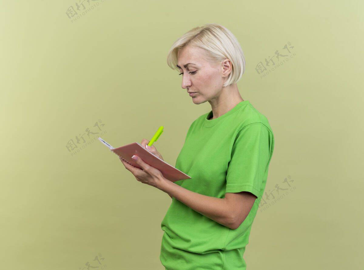 注意力专注的中年金发斯拉夫妇女站在侧视图上用橄榄绿的墙和复印空间隔离的笔在笔记本上写字个人资料笔脸