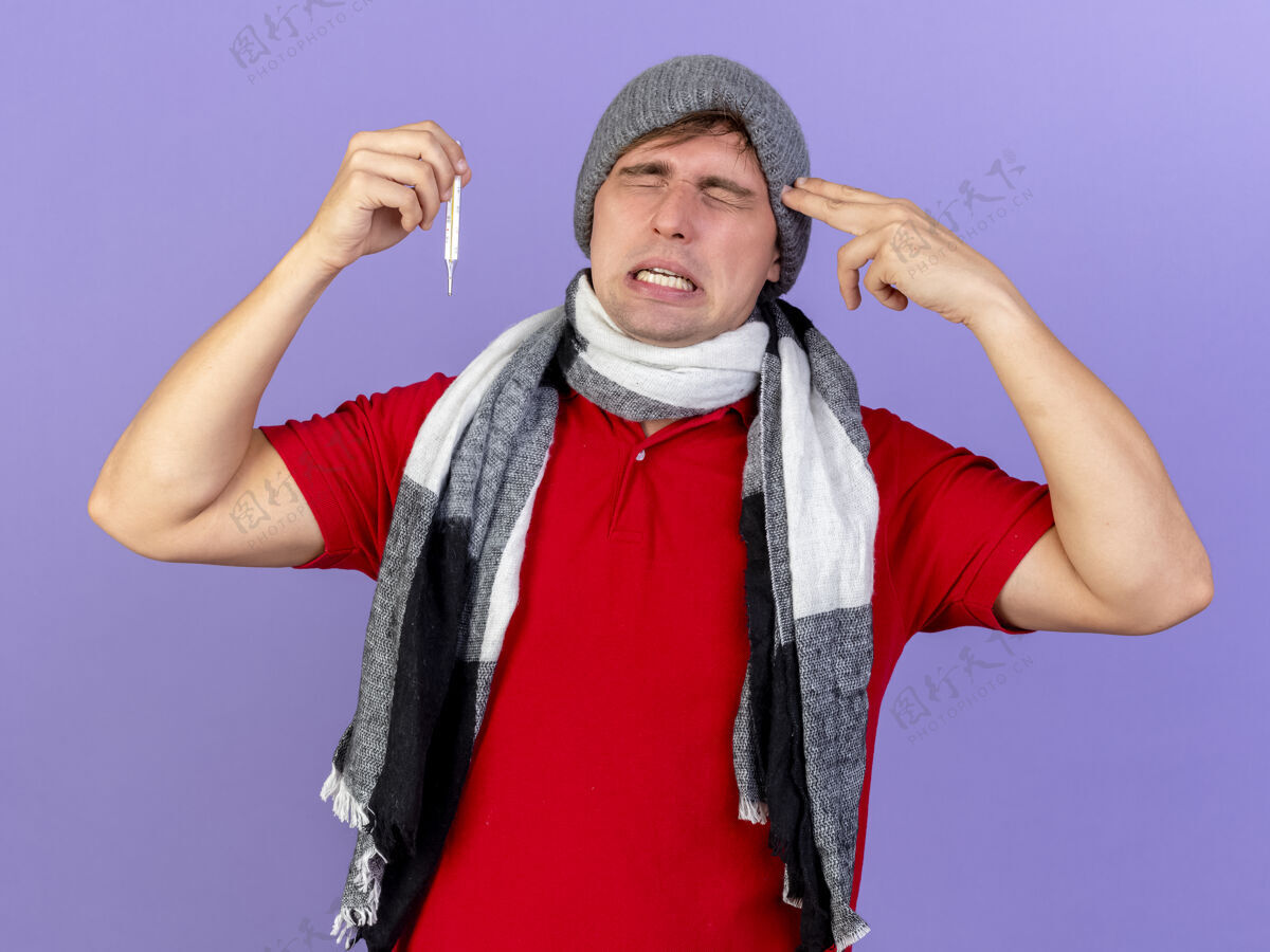 人受够了年轻英俊的金发病患戴着冬天的帽子和围巾拿着温度计做自杀手势闭着眼睛孤立在紫色的墙上摆姿势闭上围巾