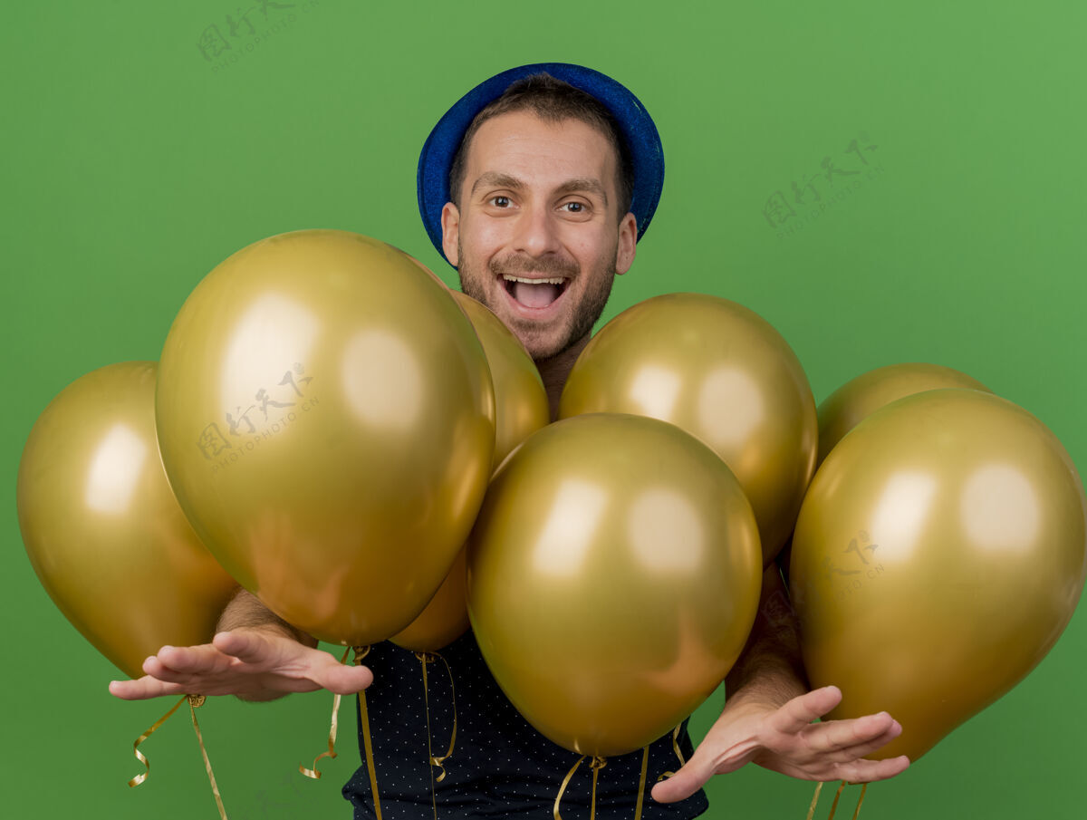人戴着蓝色派对帽的快乐帅哥站在有复制空间的绿色墙上 手上伸出氦气球帅气姿势市民