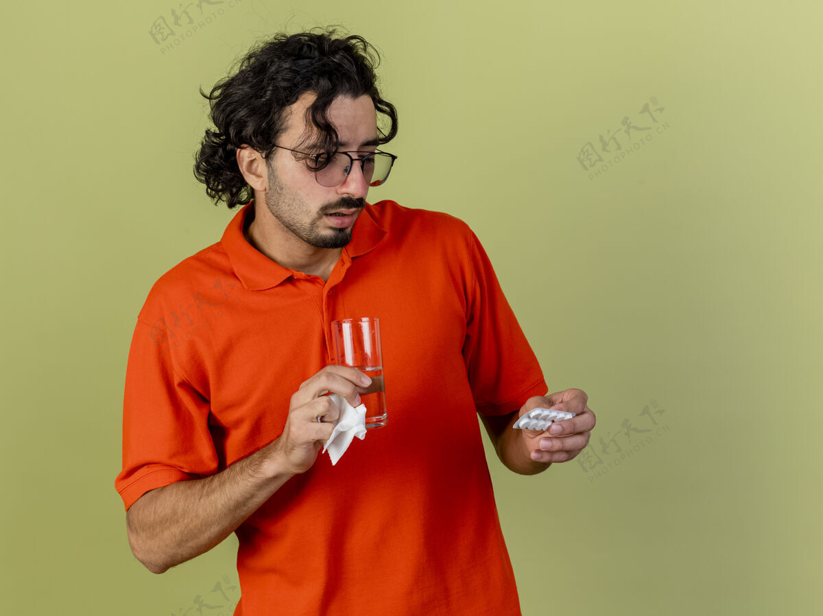 印象印象深刻的年轻人戴着眼镜拿着水杯装着药片和餐巾纸看着药片隔离在橄榄绿的墙上 留着复印空间拿着脸包装