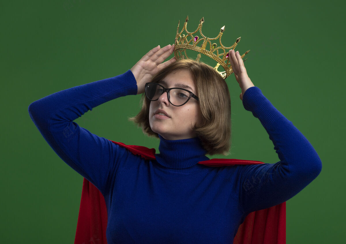 表情自信的金发女超人戴着红斗篷 戴着眼镜 望着头上戴着皇冠的一面 隔离在绿色的墙上脸公民站着