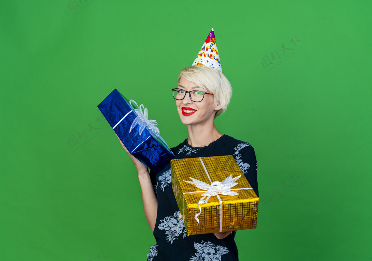 伸展微笑着的金发派对女郎戴着眼镜 戴着生日帽 拿着礼品盒 朝前伸展 看着隔离在绿色墙壁上的前面 还有复制空间人眼镜穿着