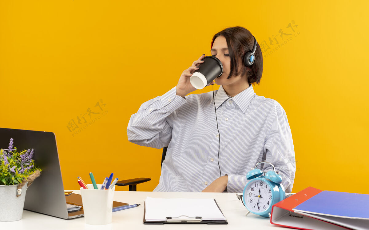 杯子年轻的呼叫中心女孩戴着耳机坐在办公桌旁 手里拿着工作工具 用塑料咖啡杯喝着咖啡 闭着眼睛隔离在橙色背景上电话女孩耳机