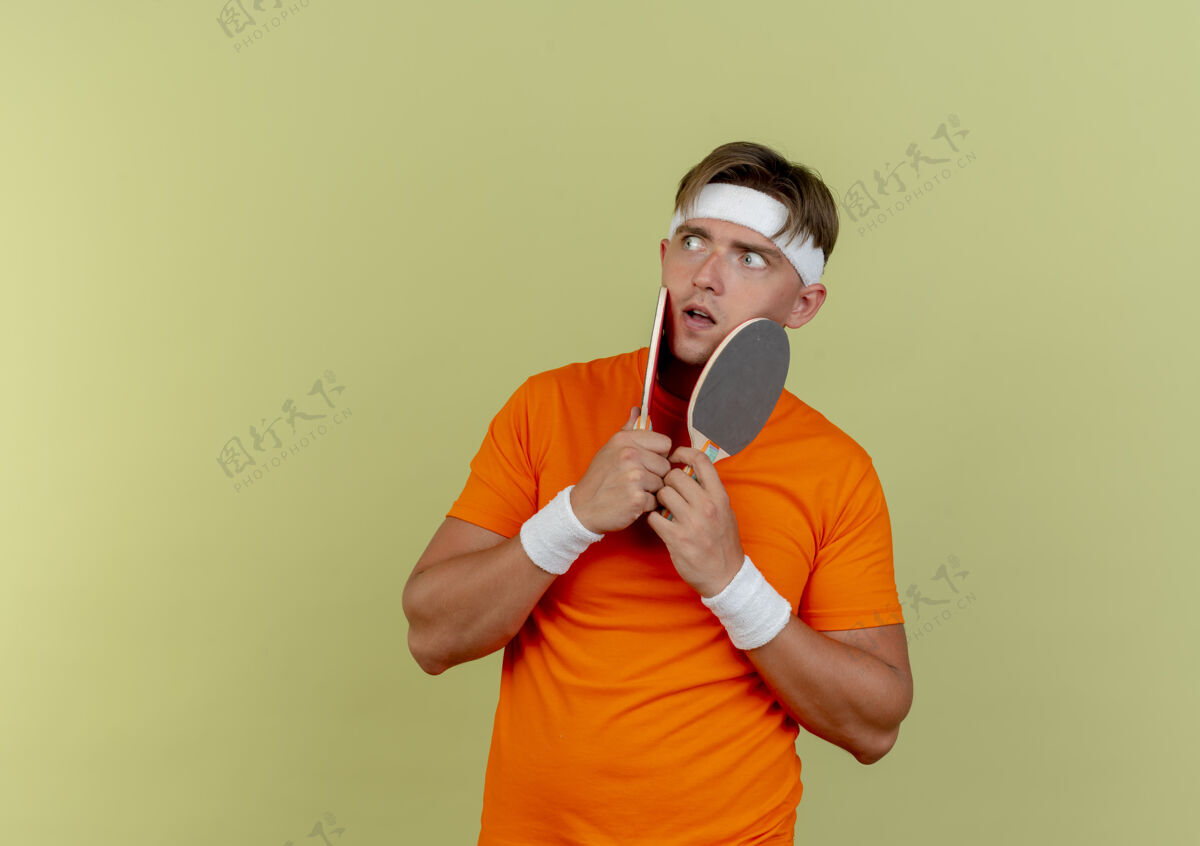 脸令人印象深刻的年轻英俊的运动男子戴着头带和腕带举行乒乓球拍和触摸他们的脸看着橄榄绿孤立的一面腕带触摸壁板
