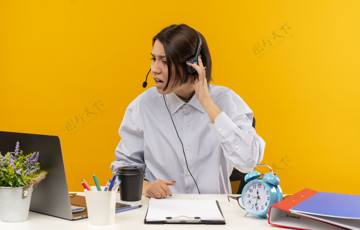电话未出租的年轻呼叫中心女孩戴着耳机坐在办公桌旁 手里拿着工作工具看着笔记本电脑 手戴着耳机隔离在橙色背景上工具笔记本电脑中心