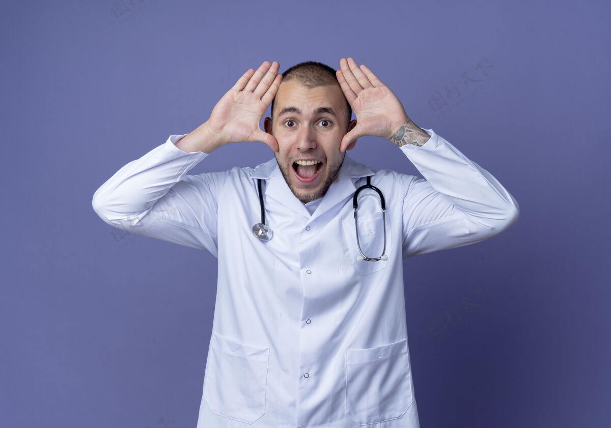 紫色令人印象深刻的年轻男医生穿着医用长袍 脖子上戴着听诊器 双手靠近头部 与紫色隔离医疗靠近手