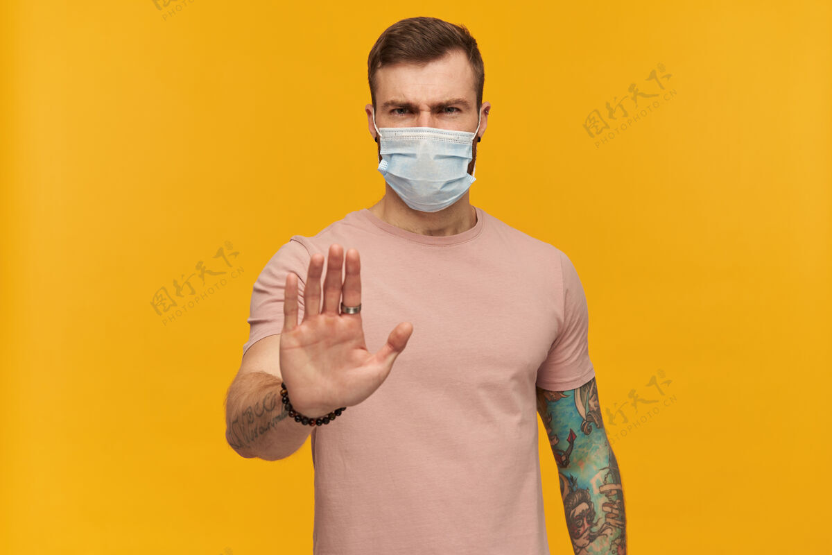 情绪一个强壮的年轻人 穿着粉色T恤 脸上戴着防冠状病毒的面具 留着胡须 纹身 手在黄色的墙上做着停止的手势年轻禁止流行