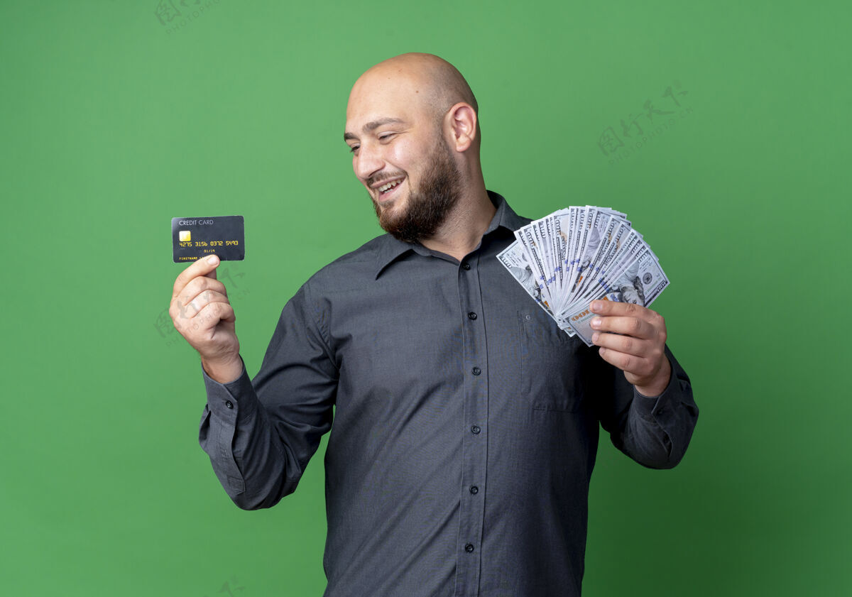 绿色快乐的年轻秃头呼叫中心男子拿着钱和信用卡看着卡上的绿色隔离信用呼叫快乐