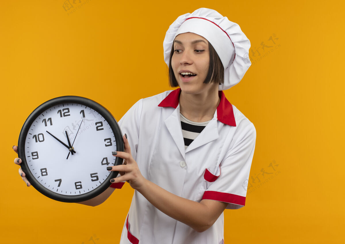制服快乐的年轻女厨师穿着厨师制服 拿着桔子看着孤立的时钟快乐厨师钟