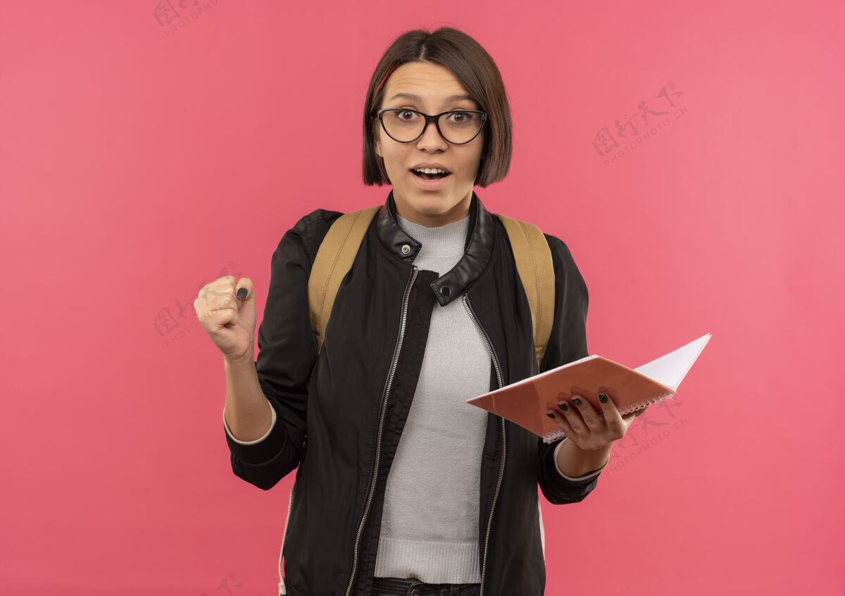 握紧印象深刻的年轻女生戴着眼镜背着书包拿着笔记本握紧拳头孤立在粉色上拳头笔记包
