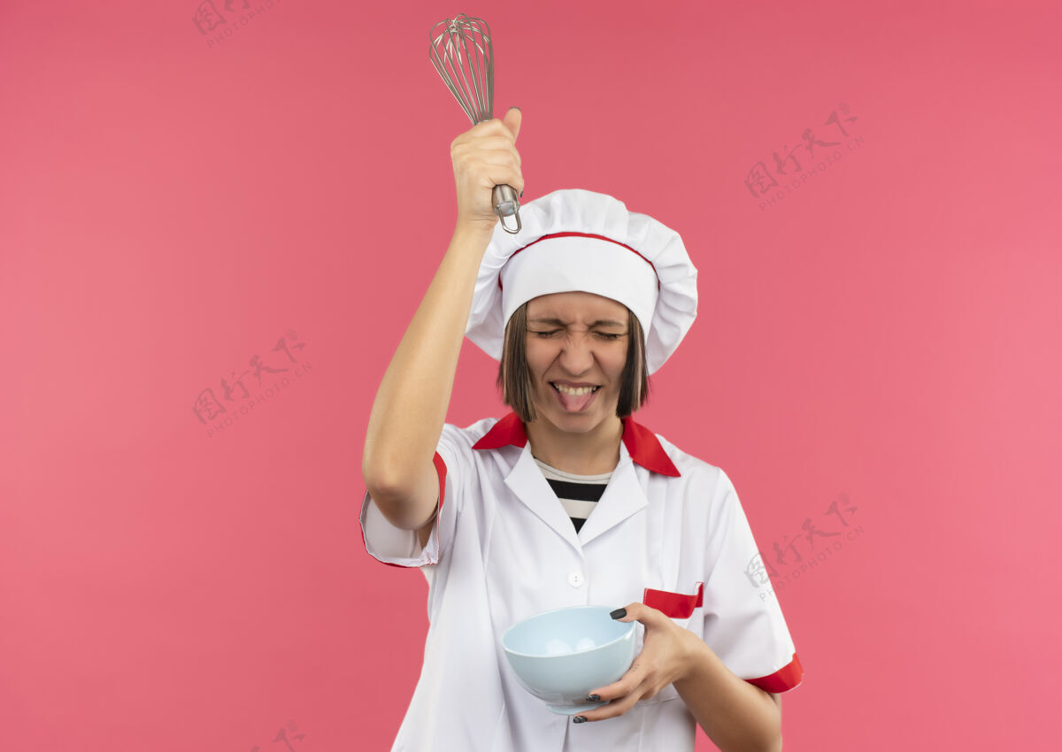 女性穿着厨师制服的顽皮的年轻女厨师拿着碗 把搅拌器举过头顶 闭着眼睛露出舌头 与粉色背景隔离 留有复印空间厨师显示拂