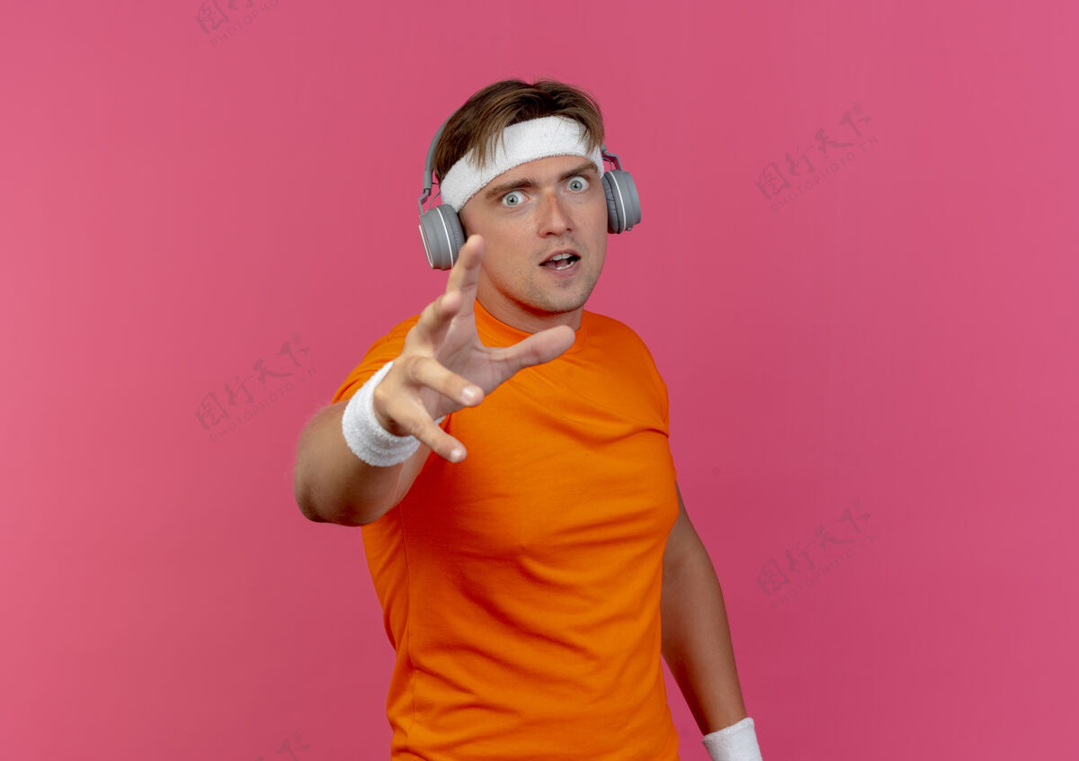 年轻令人印象深刻的年轻英俊的运动男子戴着头带和腕带和耳机伸出手孤立在粉红色腕带佩戴粉色