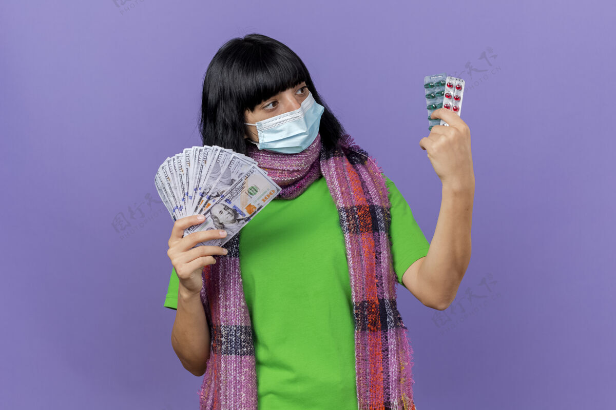 公民年轻的生病妇女戴着口罩和围巾 手里拿着钱和一包包的胶囊 看着隔离在紫色墙上的胶囊和复印空间站围巾感情