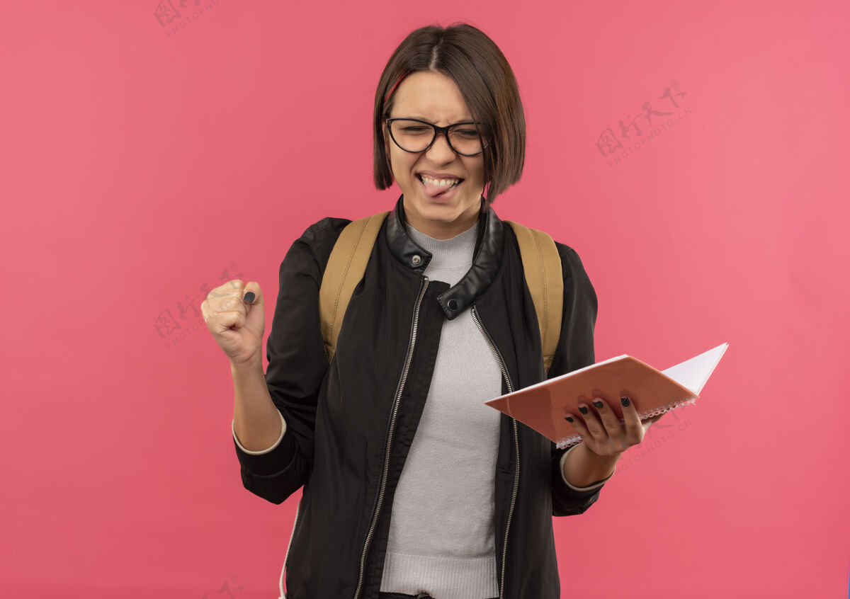 学生顽皮的年轻女学生戴着眼镜 背着袋子拿着记事本 舌头紧握着拳头 闭着眼睛 在粉色背景上 留着复印空间拿着垫年轻