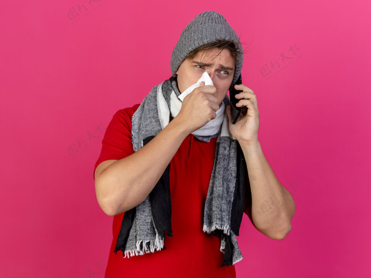 男人年轻英俊的金发病夫戴着冬天的帽子和围巾打电话用餐巾纸擦鼻子看着粉色墙壁上孤立的一面有复印空间人人疾病