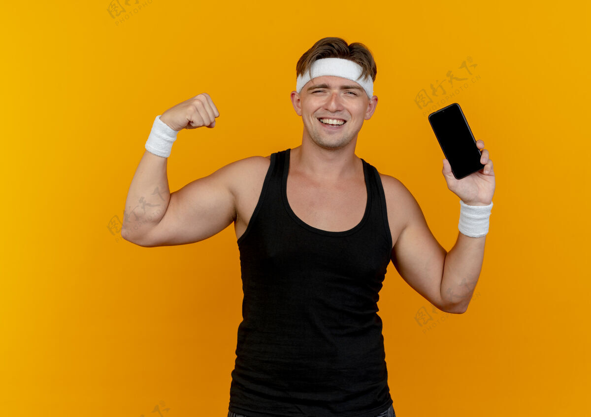 男士快乐的年轻帅气的运动型男人戴着头带和腕带拿着手机 在橙色上做着强烈的孤立的手势橙色腕带佩戴