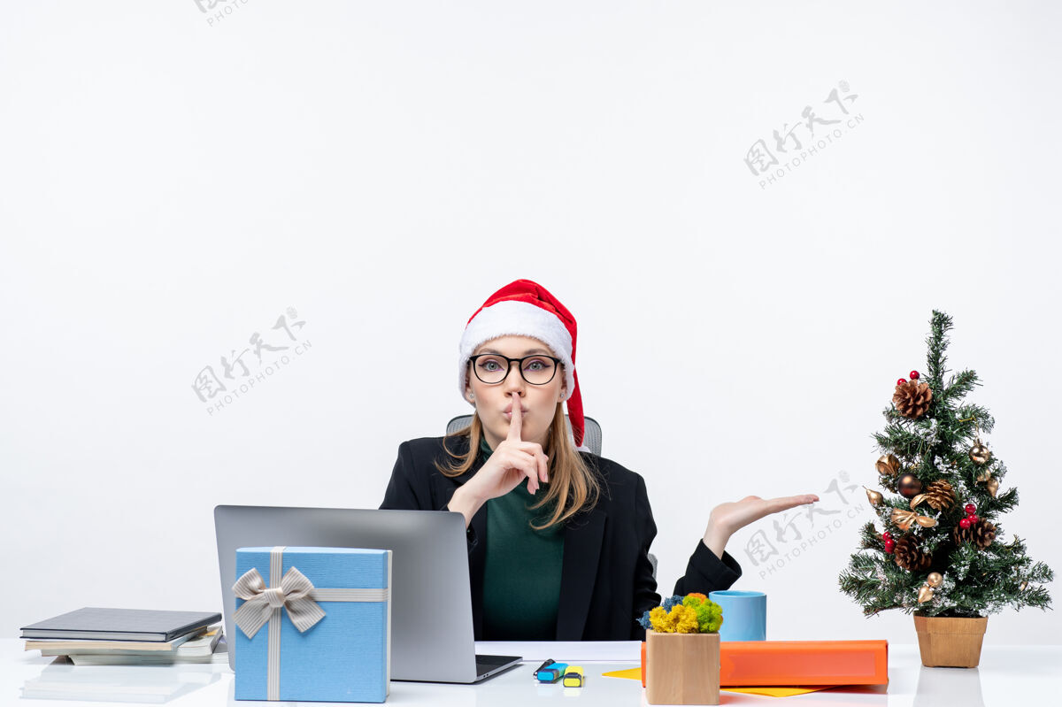 制造一个戴着圣诞老人帽子的女商人坐在一张桌子旁 桌上放着圣诞树和礼物 指着左边的东西 在办公室里做着沉默的手势电脑帽子手势