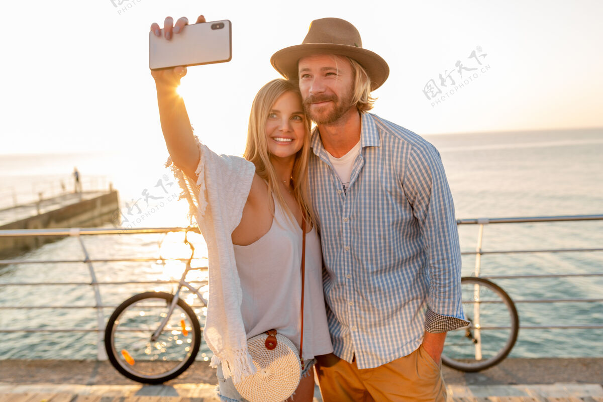 旅行者年轻迷人的笑容可掬的快乐男女骑着自行车在手机摄像头上自拍 夕阳下海边的浪漫情侣 波西米亚时髦的装扮 朋友们在一起玩得开心电话女孩大海