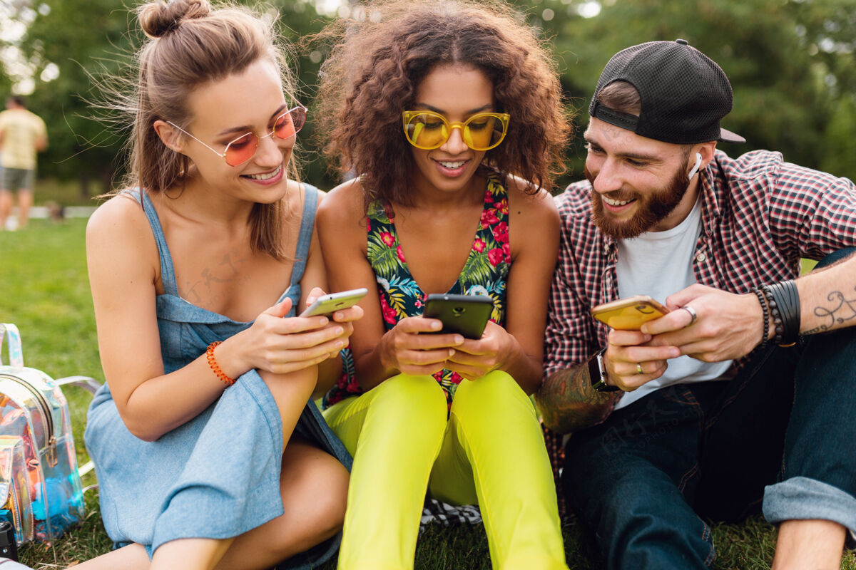 多民族快乐的年轻朋友们坐在公园里用智能手机微笑 男人和女人一起玩朋友多样性时尚