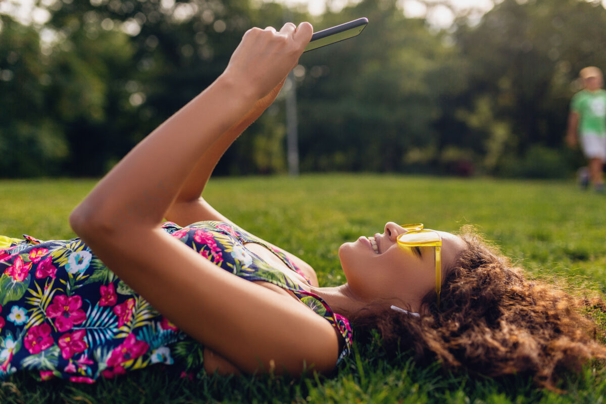 卷发年轻时尚笑脸的黑人女子用智能手机听音乐无线耳机在公园里玩得开心 夏日时尚缤纷风格 躺在草地上 戴着黄色太阳镜耳机酷休闲