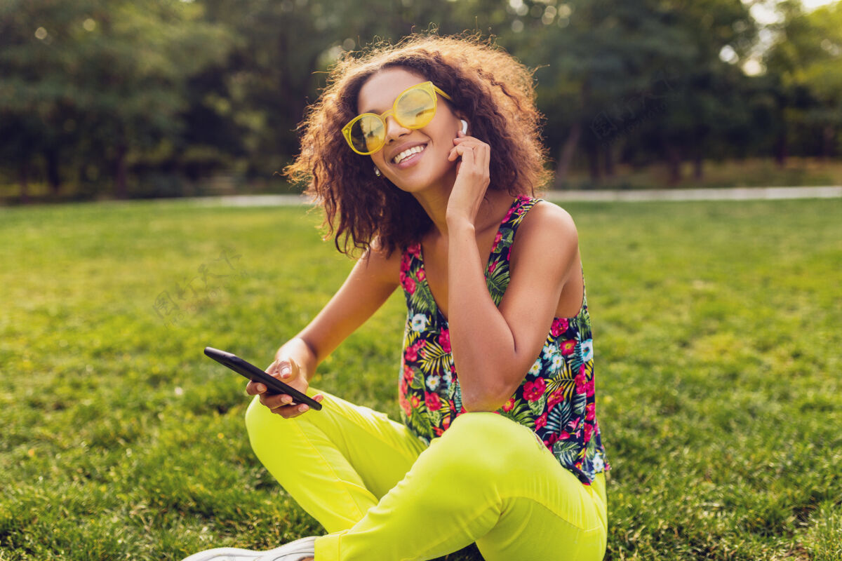 无线年轻时尚微笑的黑人女子使用智能手机 通过无线耳机听音乐 在公园里玩得很开心非裔美国人兴奋耳机