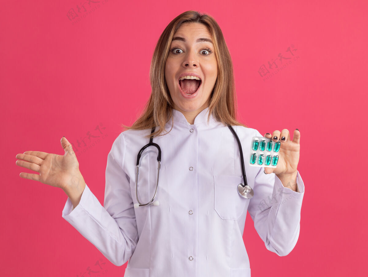 惊喜惊讶的年轻女医生穿着医用长袍 听诊器拿着药片 手被隔离在粉红色的墙上医生传播医疗