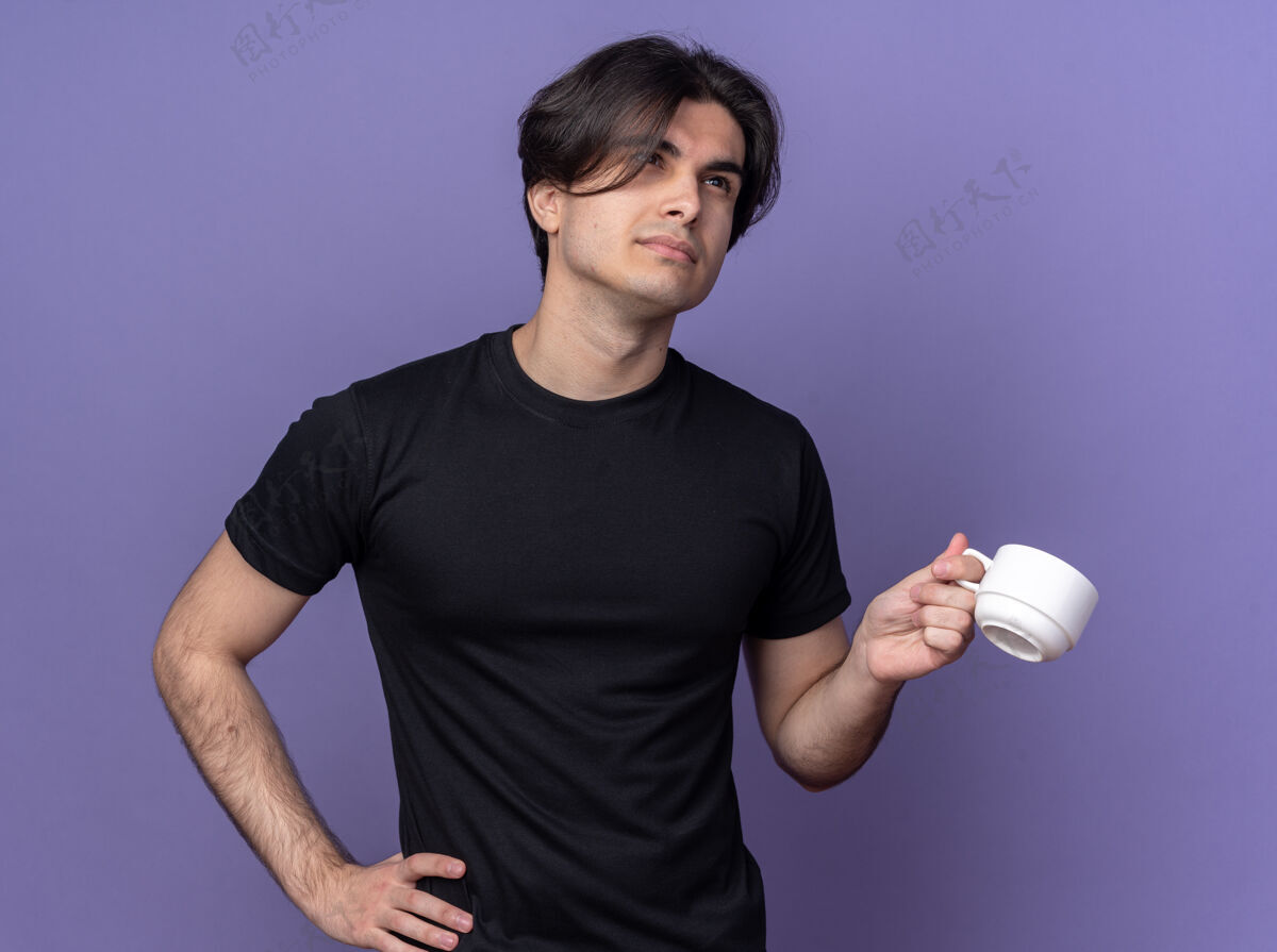 咖啡印象深刻的年轻帅哥穿着黑色t恤拿着一杯咖啡把手放在臀部隔离在紫色的墙上臀部抱着年轻