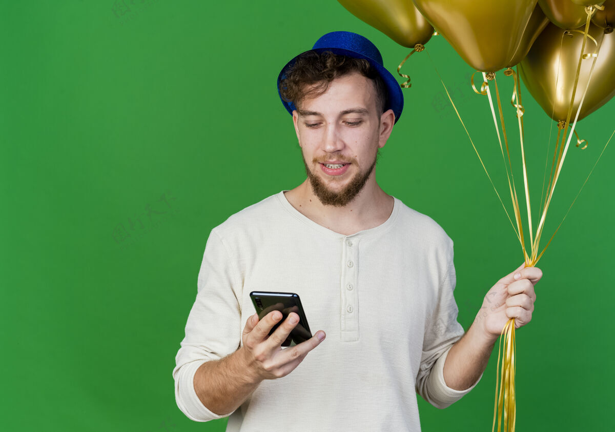 拿着高兴的年轻英俊的斯拉夫党家伙戴着党的帽子拿着气球和手机看着手机在绿色背景上与复制空间隔离帽子请绿色