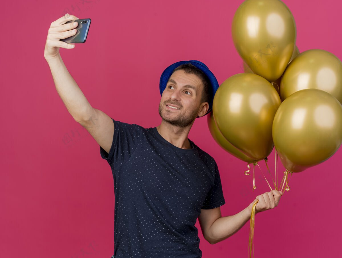 拿微笑英俊的白人男子戴着蓝色的派对帽拿着氦气球自拍看着手机在粉色背景上与复制空间隔离空间电话聚会