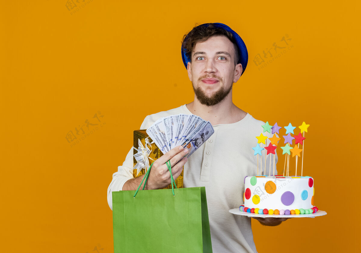 聚会年轻帅气的斯拉夫派对男 戴着派对帽 拿着礼盒 钱 纸袋和生日蛋糕 看着星星 在橙色背景下 用复制空间隔离的相机斯拉夫人星星生日