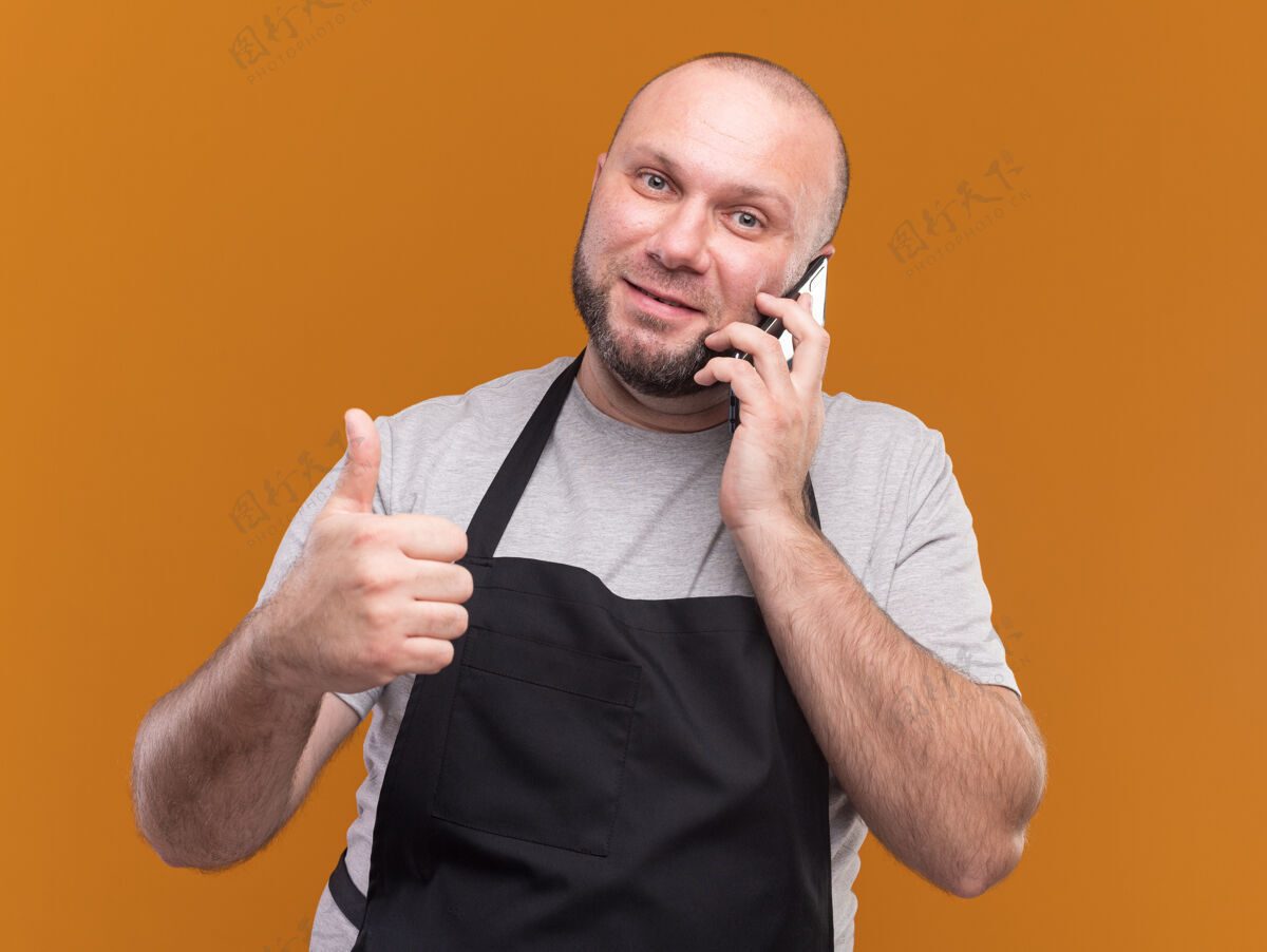 中年人高兴的斯拉夫中年男理发师穿着制服在电话里说 显示拇指向上孤立的橙色墙上男制服说话