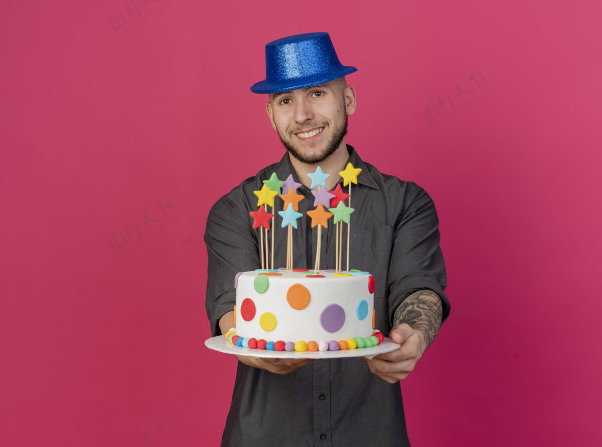 微笑面带微笑的年轻帅哥斯拉夫党人戴着党帽子伸展生日蛋糕与明星对着镜头看着摄像机在深红的背景与复制空间隔离帽子蛋糕斯拉夫