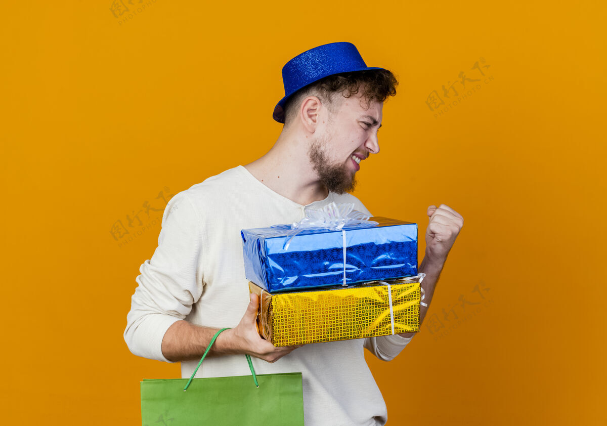 欢乐快乐的年轻英俊的斯拉夫党人戴着党的帽子拿着纸袋和礼品盒看在一边做是的手势孤立在橙色背景与复制空间聚会盒子拿着