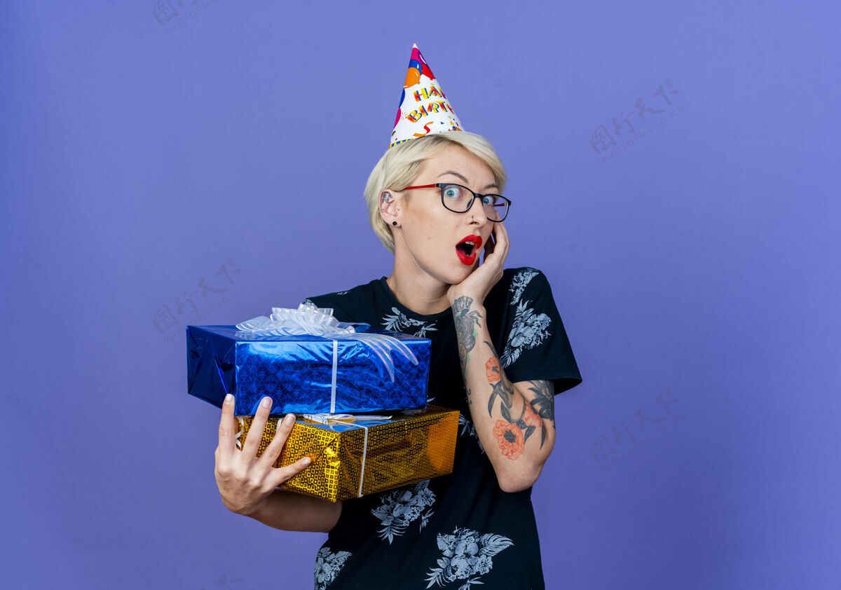 空间惊讶的年轻金发派对女孩戴着眼镜 戴着生日帽 拿着礼品盒 看着相机 手放在脸上 紫色背景上 留着复制空间拿着眼镜帽子