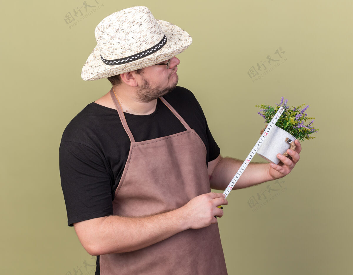 思想年轻的男园丁戴着园艺帽 用卷尺在橄榄绿的墙上测量花盆里的花测量花园艺