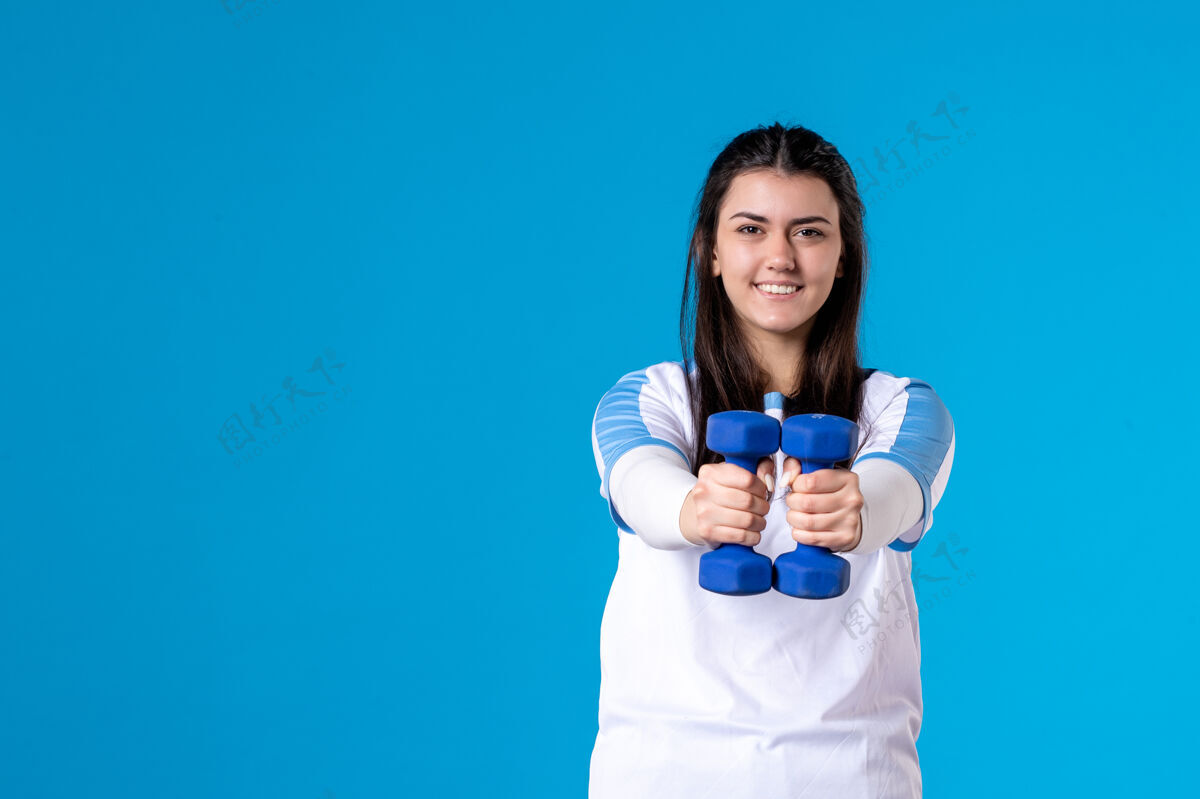 锻炼正面图：年轻女子手持蓝色哑铃站在蓝色墙上获胜者正面运动