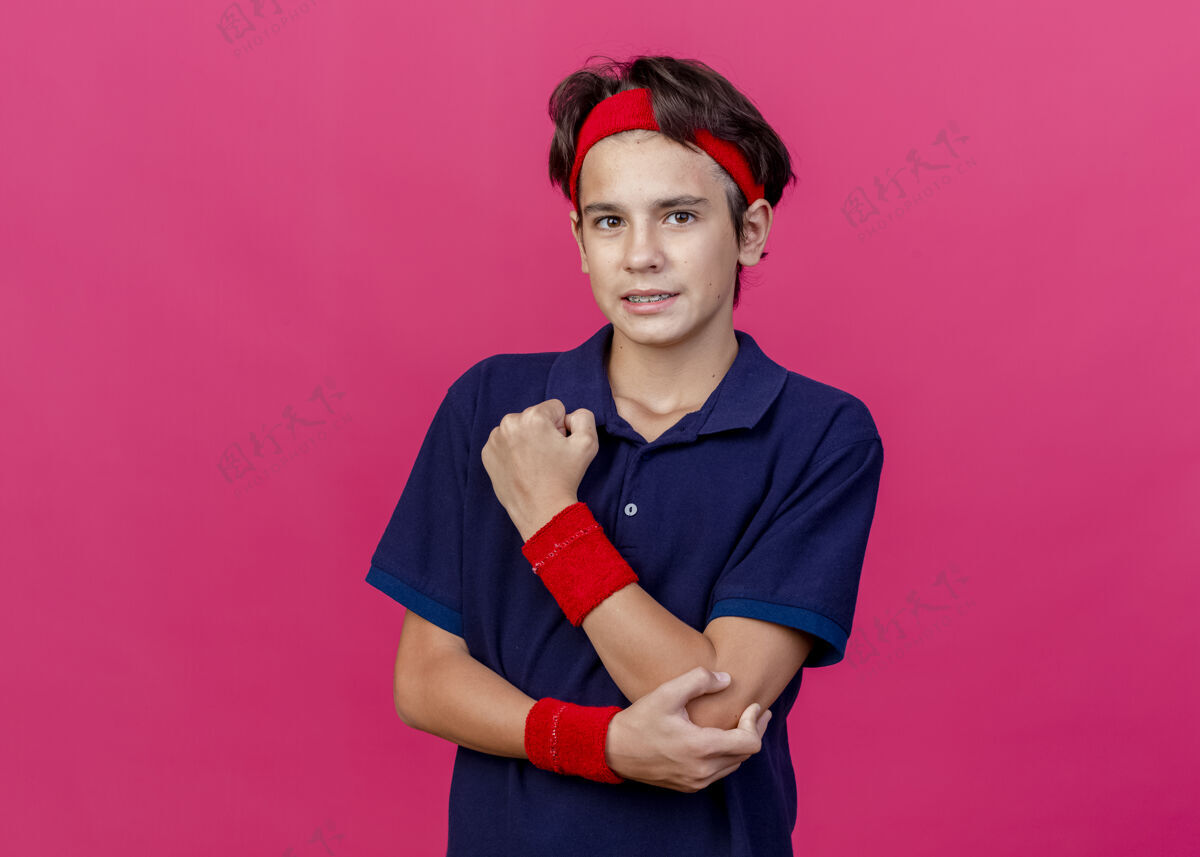运动疼痛的年轻英俊的运动男孩戴着头带和护腕 戴着牙套 手放在手肘上 看着被隔离在深红色背景上的相机 还有复制空间背带肘部疼痛
