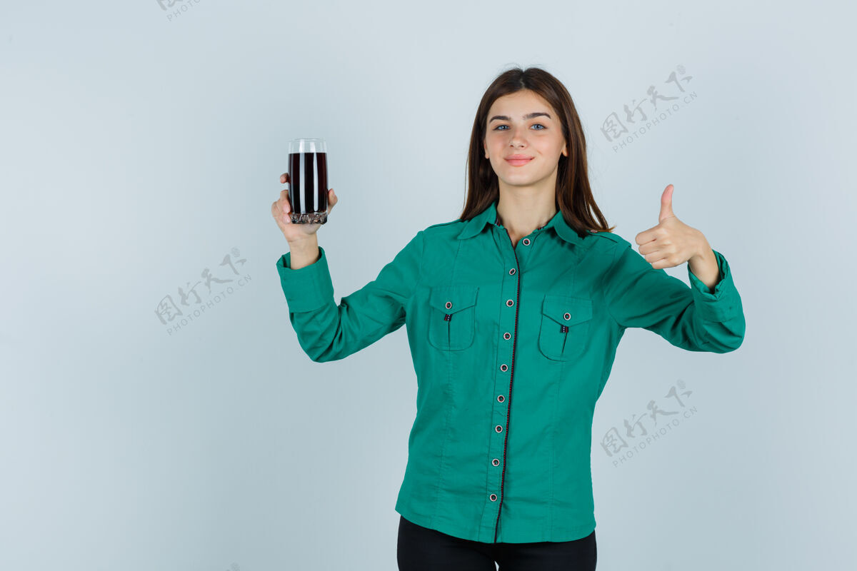 玻璃年轻女孩拿着一杯黑色液体 穿着绿色上衣 黑色裤子 竖起大拇指 看起来很开心正面图青年前面女性